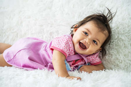 Childcare Concept. Portrait of cute little mixed race baby girl smiling and lying on white bed sheets at home. infant child crawling on bed in the bedroom. Selective focusの写真素材
