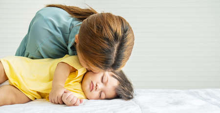 Young mother is kissing her cute little baby who is sleeping in bed at home, Mom and daughter relax in a white bedroom. Family at home. Love, trust and tenderness concept.の写真素材