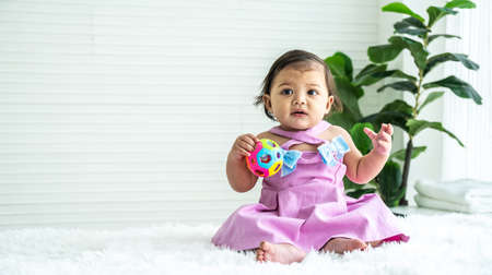 Adorable mixed race baby girl in pink clothes sitting on bed at home, looking away, playing toy color ball. Kid at home and childhood conceptの写真素材