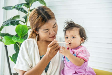 Mother feeding her baby daughter with spoon. Mother giving healthy food to her adorable child at home, Baby food.の写真素材