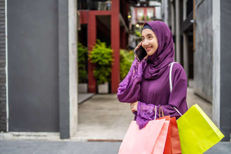 Modern lifestyle of arab muslim young woman in veil hijab holding shopping bags walking on the city street. while talking on mobile phoneの写真素材