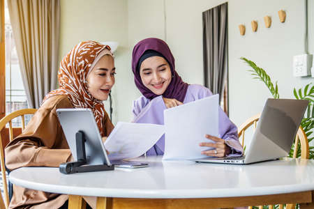 Two Asian muslim women wearing hijab working together with using laptop pc tablet and paper showing graphic chart in living room at home, respect and collaboration concept.の写真素材