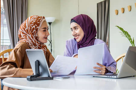 Two Asian muslim women wearing hijab working together with using laptop pc tablet and paper showing graphic chart in living room at home, respect and collaboration concept.の写真素材