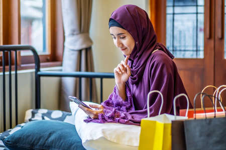 Online shopping concept,Young asian muslim woman sitting on couch in home living room. holding smartphone making online shopping and many shopping bag, browsing online sale website on cellphoneの写真素材