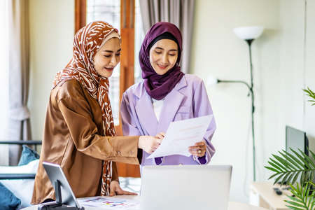 Two Asian muslim women wearing hijab working together with using laptop pc tablet and paper showing graphic chart in living room at home, respect and collaboration concept.の写真素材