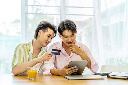 Portrait of young gay couple, lgbt man holding credit card enjoy shopping online with tablet or laptop at home. LGBT and love concept.の写真素材