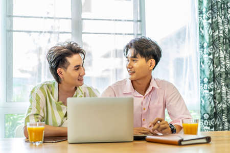 Asian businessman gay work with smile and laugh in office. Attractive male lgbtq collegues working together with using laptop pc and paper showing graphic chart in living room, Lgbt love conceptの写真素材