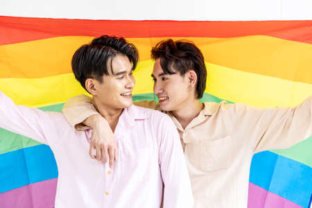 Portrait of Happy gay couple showing their love with rainbow flag. holding each other and holding up an LGBTQ flag, They are laughing, LGBT and love concept.の写真素材
