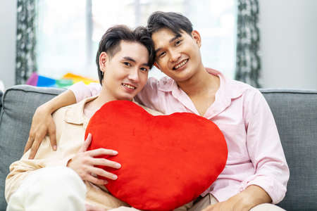 Valentine's Day. Young attractive gay couple sitting on sofa at home look at camera and holding red heart pillow together with love and understanding. Concept of LGBT and Loveの写真素材