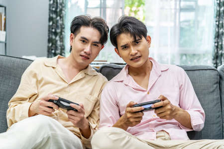 Two young males relax on weekend sit on couch enjoy console video game push buttons on controllers. Two Male Players spend free time together play online game having funの写真素材