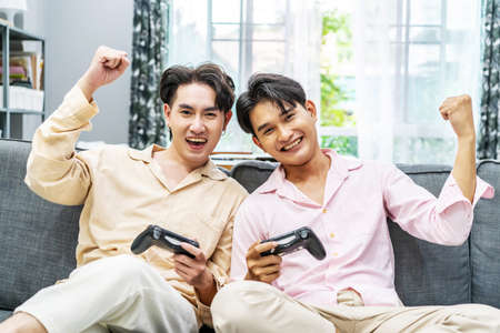 Two young males relax on weekend sit on couch enjoy console video game push buttons on controllers. Two Male Players spend free time together play online game having funの写真素材
