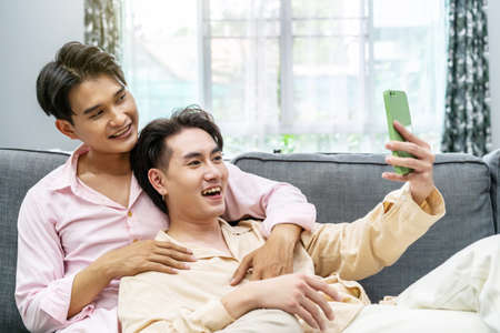 Guys Smile and Laugh in Love. Cute Male Homosexual Gay Couple Spend Time Together, Men Taking Selfie on Smartphone, sitting on a sofa in sitting room, LGBTQ, LGBT Pride Month, Gay Pride Symbolの写真素材