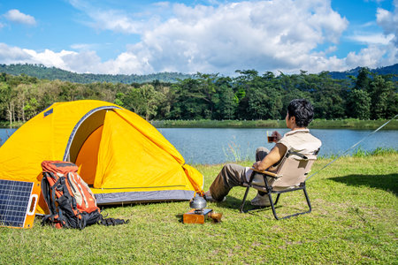 Young Asian traveling man relax and enjoy outdoor lifestyle camping in forest mountain on summer holiday travel vacation. Attractive male drinking coffee while enjoys drink among green treesの写真素材