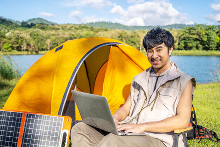 Young Asian traveling man relax and enjoy outdoor lifestyle camping in forest mountain on summer holiday. Attractive male relaxing in chair while working on laptop during camping day in natureの写真素材