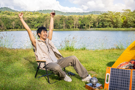 Young Asian traveling man relax and enjoy outdoor lifestyle camping in forest mountain on summer holiday. Attractive male drinking coffee and raise your hands refreshingly in the midst of natureの写真素材