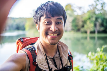 Young asian attractive male tourists holding camera mobile phone and taking selfie on green summer lawn while looking at camera, admiring the beauty natureの写真素材