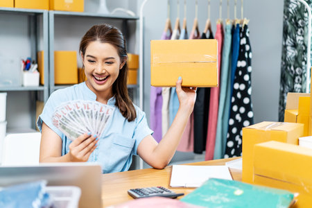 Entrepreneurs Small Business SME freelance, young Asian woman holding US dollars lot of money with box, she is happy and proud smile work at home, online marketing packing box delivery, online salesの写真素材