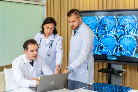 Team of three doctors discusses the results of an MRI scan of the patient's head in the consulting room of modern hospital, physician discussing about treatment for patient looking in laptopの写真素材