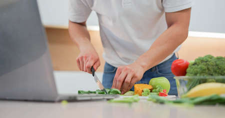 close up man hand using knife to cut vegetables on board and preparing food while open laptop to...