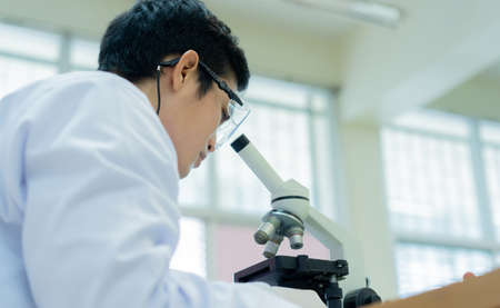 close up rear view of young asian scientist looking at microscope to research or record and analyze about scientific experiment result at laboratory for healthcare and pharmaceutical conceptの写真素材