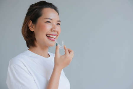 close up young asian woman smiling with hand holding dental aligner retainer (invisible) on gray background of dental clinic for beautiful teeth treatment course conceptの写真素材