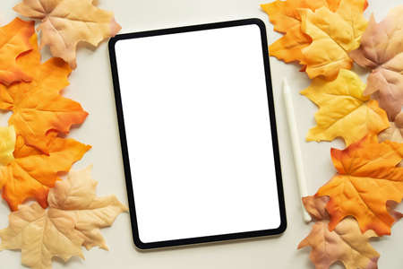 close up top view mockup of white blank digital tablet screen and stylus pen with group of dried orange color maple leaves background texture or autumn season collection design conceptの写真素材