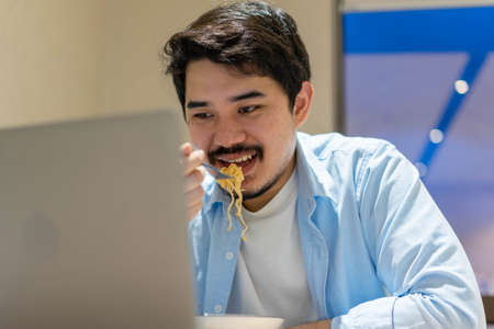 close up young middle eastern man eating noodle ramen during check work on laptop at office in the late night for overwork lifestyle conceptの写真素材