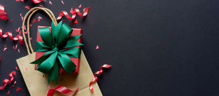 close up top view of christmas gift box with shopping bag with red rolling ribbon and confetti on black background with copy space for black Friday season and happy new year festival concept