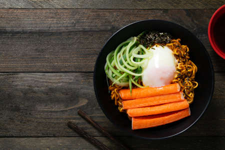 Spicy instant noodle with crab stick and onsen egg and cucumber in black bowl on wooden table.の写真素材