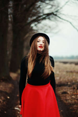 Beautiful young girl with long straight hair, light make-up in a stylish trendy in a black jacket and red leather skirt in black hat outdoors in a parkの写真素材