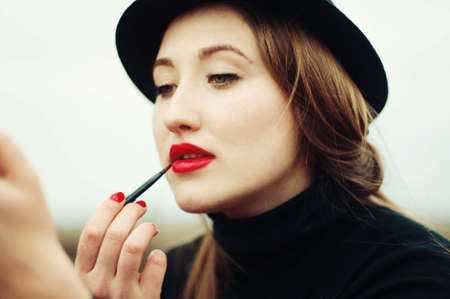 Beautiful young girl with long straight hair, light make-up in a stylish trendy in a black jacket and red leather skirt in black hat outdoors in a park on a background of trees. The girl looks  cameraの写真素材