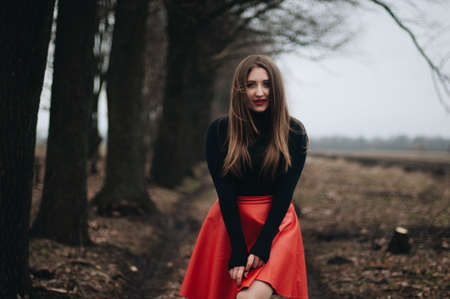 Beautiful young girl with long straight hair, light make-up in a stylish trendy in a black jacket and red leather skirt in black hat outdoors in a park on a background of trees. The girl looks  cameraの写真素材