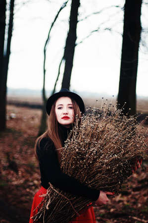 Beautiful young girl with long straight hair, light make-up in a stylish trendy in a black jacket and red leather skirt in black hat outdoors in a park on a background of trees. The girl looks  cameraの写真素材