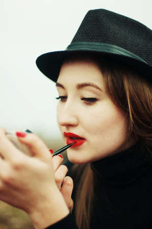 Beautiful young girl with long straight hair, light make-up in a stylish trendy in a black jacket and red leather skirt in black hat outdoors in a park on a background of trees. The girl looks  cameraの写真素材