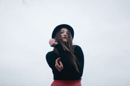 Beautiful young girl with long straight hair, light make-up in a stylish trendy in a black jacket and red leather skirt in black hat outdoors in a park on a background of trees. The girl looks  cameraの写真素材