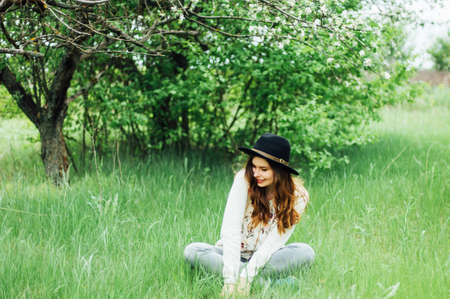 Stylish spring bohemian outfits. Wearing a white sweater and black hat in blooming garden. Beautiful young woman with long wavy hair thin slender figure perfect body and pretty face natural make-up.の写真素材