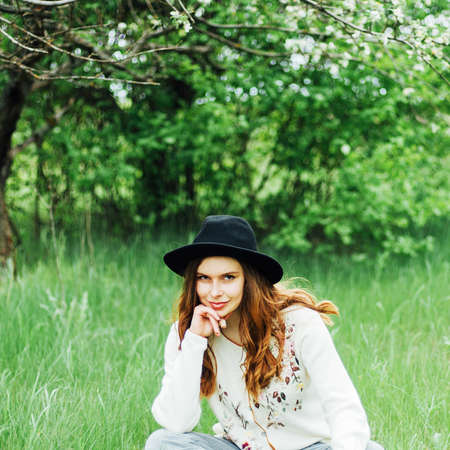 Stylish spring bohemian outfits. Wearing a white sweater and black hat in blooming garden. Beautiful young woman with long wavy hair thin slender figure perfect body and pretty face natural make-up.の写真素材
