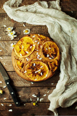 Delicious fresh homemade pie with orange zest, milk, butter, eggs. Decorated with slices of orange on a wooden background. Cake sprinkled with powdered sugar. Decorated with wild flowersの写真素材