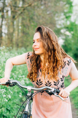 Outdoor fashion portrait of elegant lady standing near hipster bicycle in pink stylish dress. Enjoy and having fun spring day in the park with a vsco filters. Beautiful  girl with long thick wavy hairの写真素材