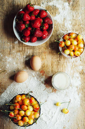 Cooking cake with fresh ingredients for preparing in cooking. Rustic dark atmosphere. Flour, eggs and fresh strawberries, cherries, butter, flour over on wooden background. Series, recipe step on stepの写真素材