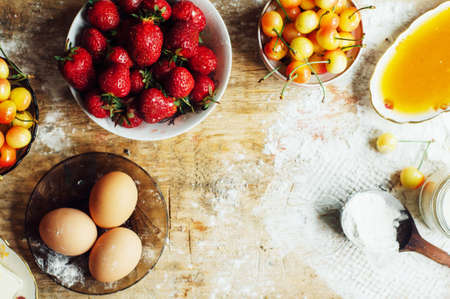 Cooking cake with fresh ingredients for preparing in cooking. Rustic dark atmosphere. Flour, eggs and fresh strawberries, cherries, butter, flour over on wooden background. Series, recipe step on stepの写真素材