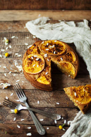 Delicious fresh homemade pie with orange zest, milk, butter, eggs. Decorated with slices of orange on a wooden background. Cake sprinkled with powdered sugar. Decorated with wild flowersの写真素材