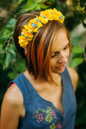 Portrait of a beautiful young woman wearing wreath. Young beautiful woman posing with flowers bouquet on her head. Girl posing in a decorative wreath of artificial flowers on outdoor in the summerの写真素材