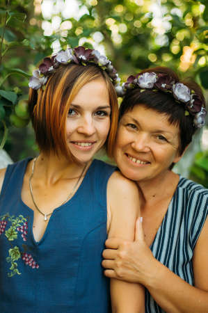 Portrait of a smiling mother and daughter are posing in wreaths of artificial flowers from the outdoors on a background of trees. The concept of family. The relationship of the mother and daughter.の写真素材