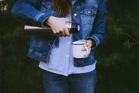 Traveler girl pouring tea from vacuum flask cup, outdoors. Young woman drinking tea at cup. Theme travel. Woman pouring a hot drink in mug from vacuum flask. Girl drinking tea during hikeの写真素材