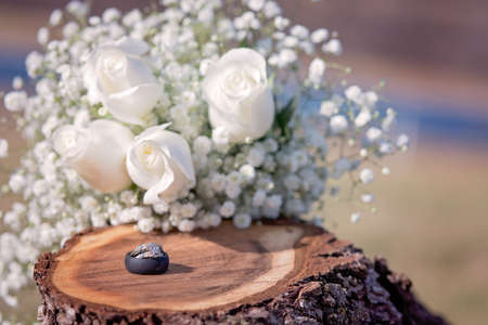 Wedding and engagement ring with black silicone mans ring on wooden stump with white roses in backgroundの写真素材