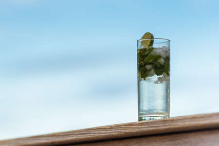 Tall clear glass with ice, mint leaves, lime slice and bubbly spirits sitting outdoors on a deck railの写真素材