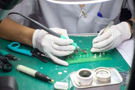 Close-up technician female on rubber gloves and work suit is using soldering to weld electronics ...