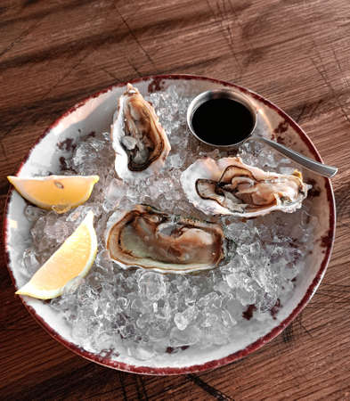 Fresh open oyster with lemon on a plate with ice and sauce on the dark kitchen table. Seafood appetizer.の写真素材