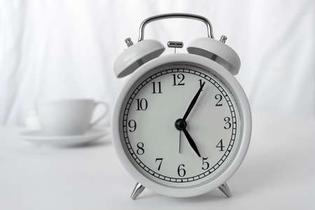 The white alarm clock is on the table.Behind it is a white cup of coffee on a light background. The...